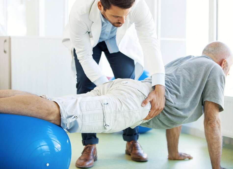 Physio helps senior man do exercises on Swiss ball