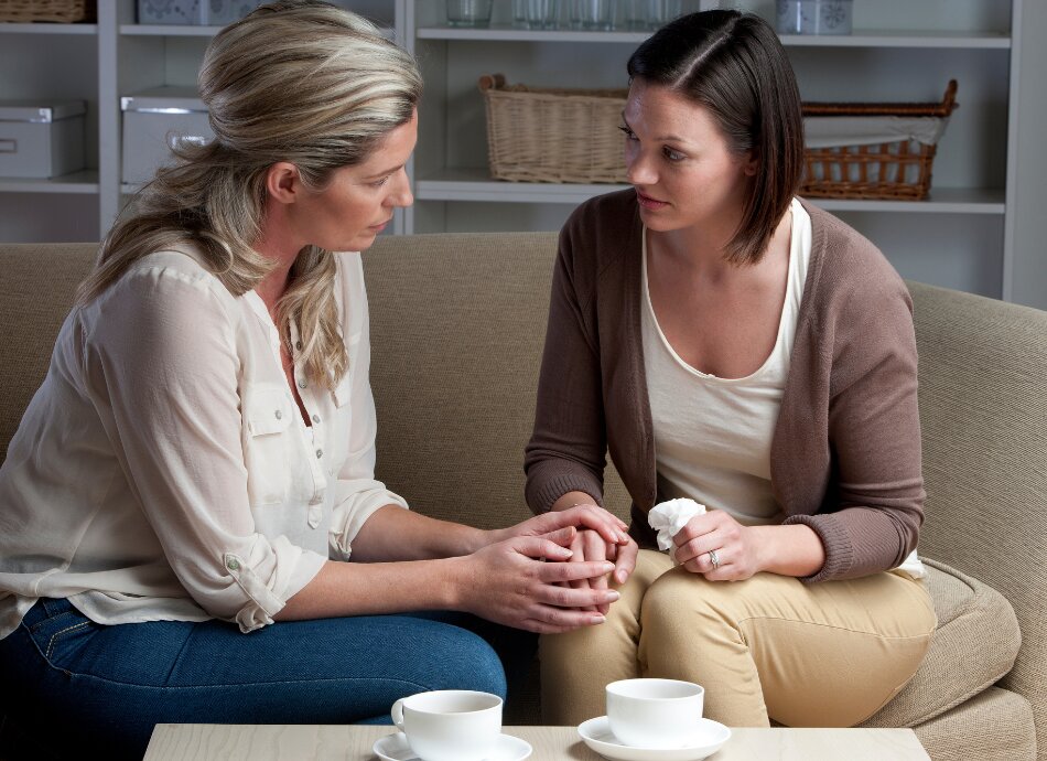 Two women sitting on couch and talking while one comforts the other