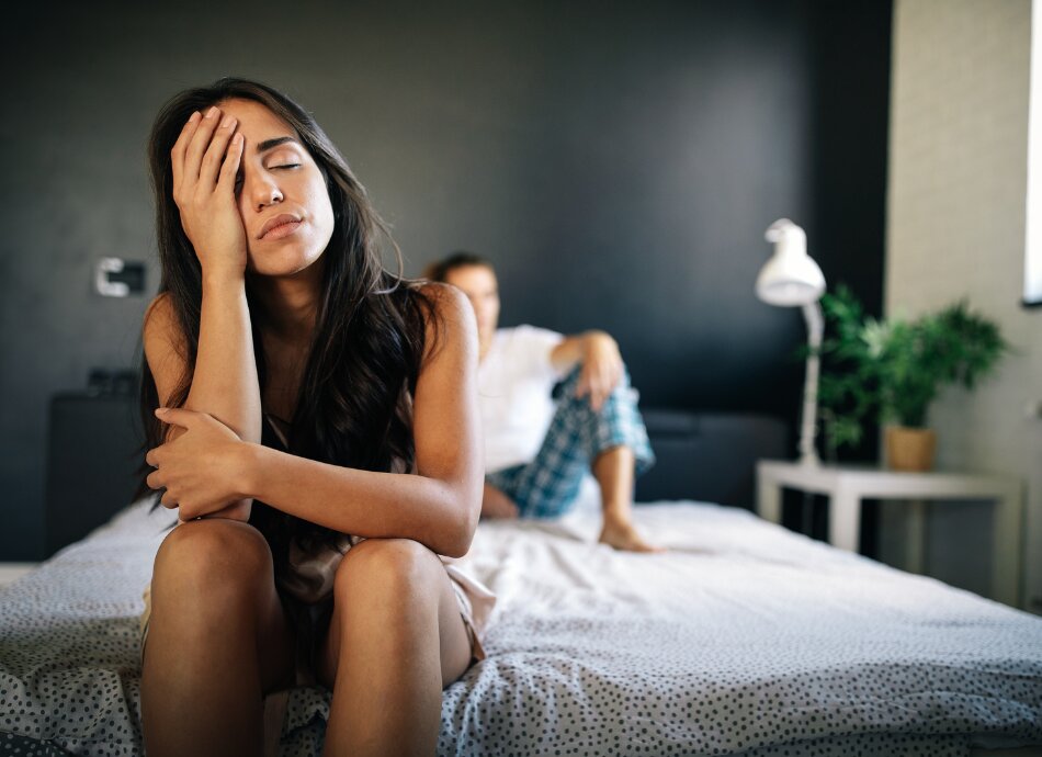 Unhappy woman on end of bed with partner blurred out behind her