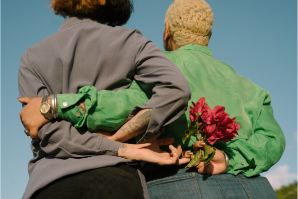 2 friends embrace and one has flowers behind her back